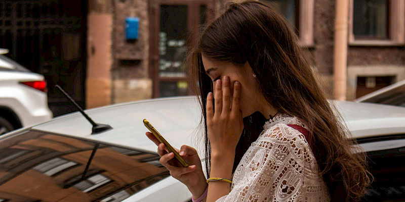 Das Bild zeigt eine junge Frau, die am Handy ist und die sich hinter einem weißen Auto befindet. Sie scheint zu warten. Gegenüber ist ein weiteres farbiges Auto zu sehen.