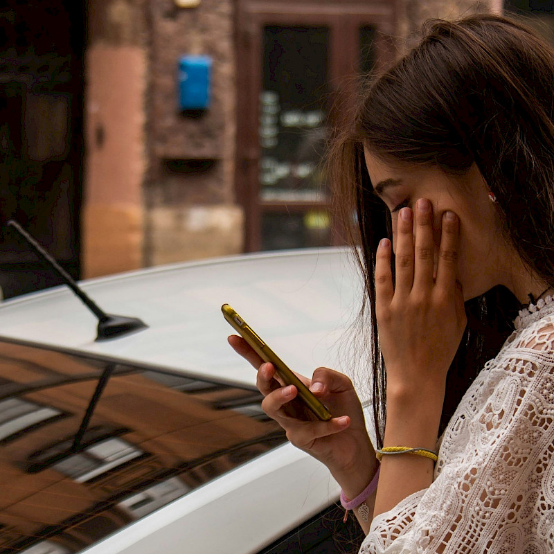 Das Bild zeigt eine junge Frau, die am Handy ist und die sich hinter einem weißen Auto befindet. Sie scheint zu warten. Gegenüber ist ein weiteres farbiges Auto zu sehen.