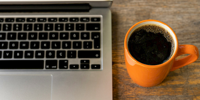 Auf dem Bild ist der AUsschnitt eines silbernen Laptops und dessen Tatstatur zu erkennen. Daneben sreht eine orangefarbige Tasse, die mit schwarzem Kaffee gefüllt ist. Der Laptop und die Tasse stehen auf einem massiven Holztisch. Der Untergrund des Tisches ist zu erkennen.