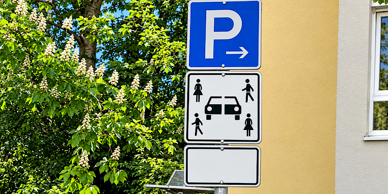 Auf dem Bild ist ein Verkehrsschild, das Carsharing anzeigt zu sehen. Das Verkehrsschild zeigt unter dem Parkenschild vier Personen (zwei männliche und zwei weibliche Piktogramme) und ein durchtrenntes Fahrzeug. Das Zeichen weist klar darauf hin, dass dieser Parkplatz Carsharing-Fahrzeugen vorbehalten ist.