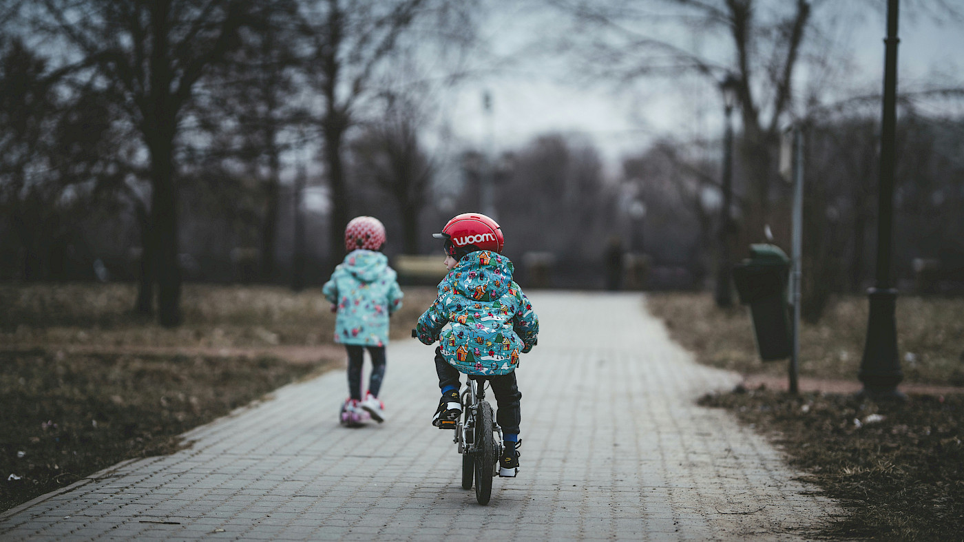 Das Bild zeigt zwei Kleinkinder, die auf einem Kinderfahrrad und einem Roller auf einem Weg, der geplfastert ist, unterwegs sind. Die Umgebung wirkt winterlich, es befinden sich kahle Bäume neben dem Weg.