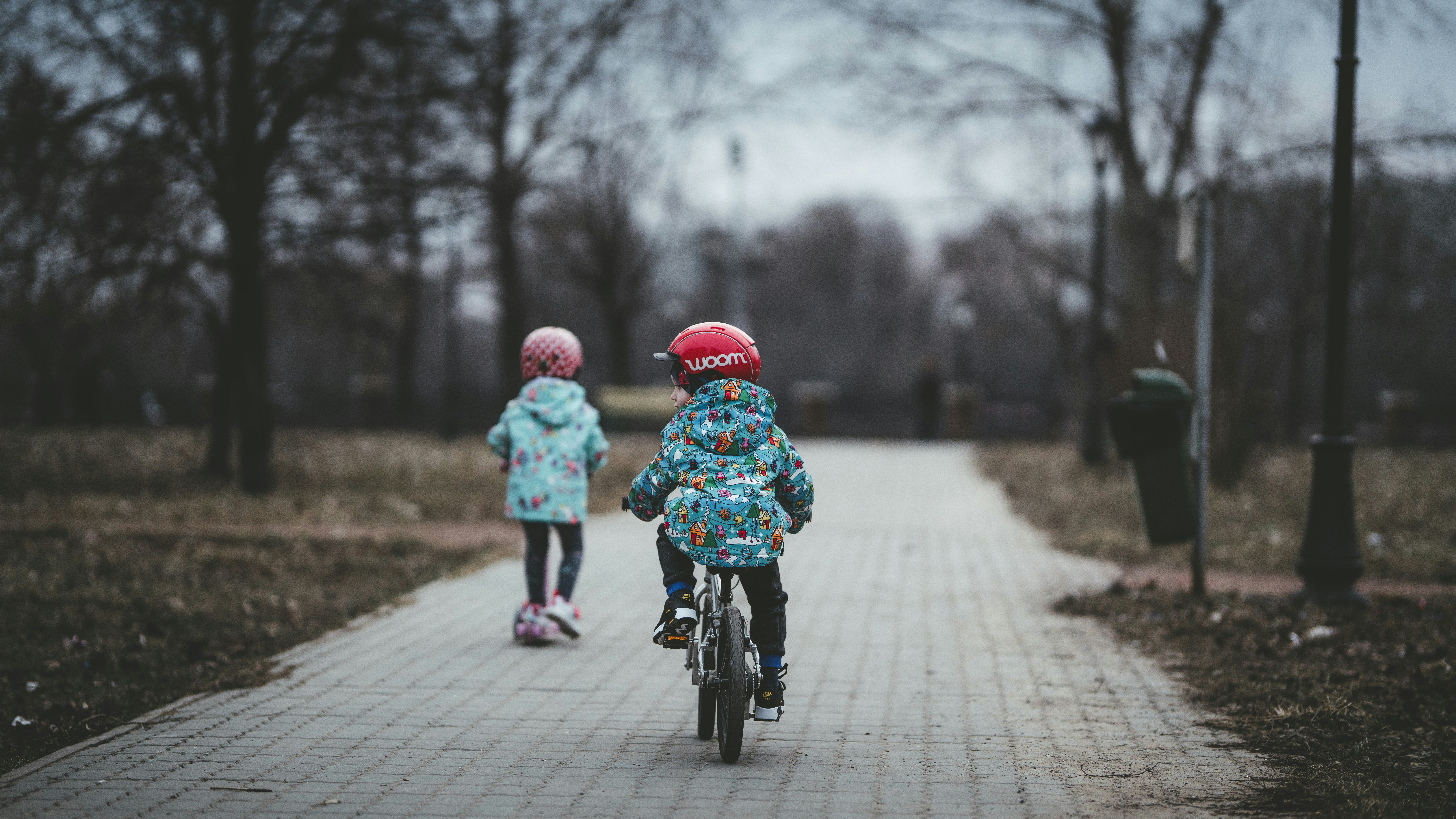 Das Bild zeigt zwei Kleinkinder, die auf einem Kinderfahrrad und einem Roller auf einem Weg, der geplfastert ist, unterwegs sind. Die Umgebung wirkt winterlich, es befinden sich kahle Bäume neben dem Weg.