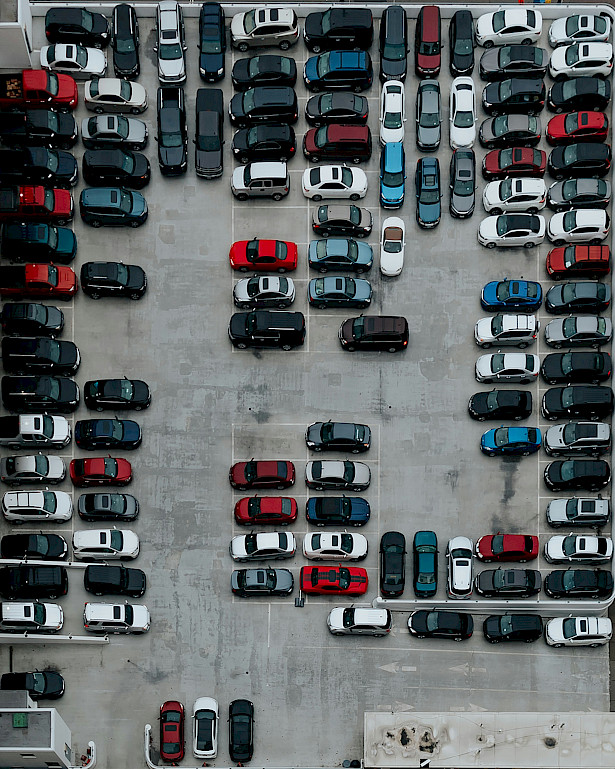 Das Bild zeigt eine Straße, im Hintergrund befinden sich Häuser. An den Seitenrändern der Straße stehen viele parkende Autos. Die Autos weisen verschiedene Farben auf.