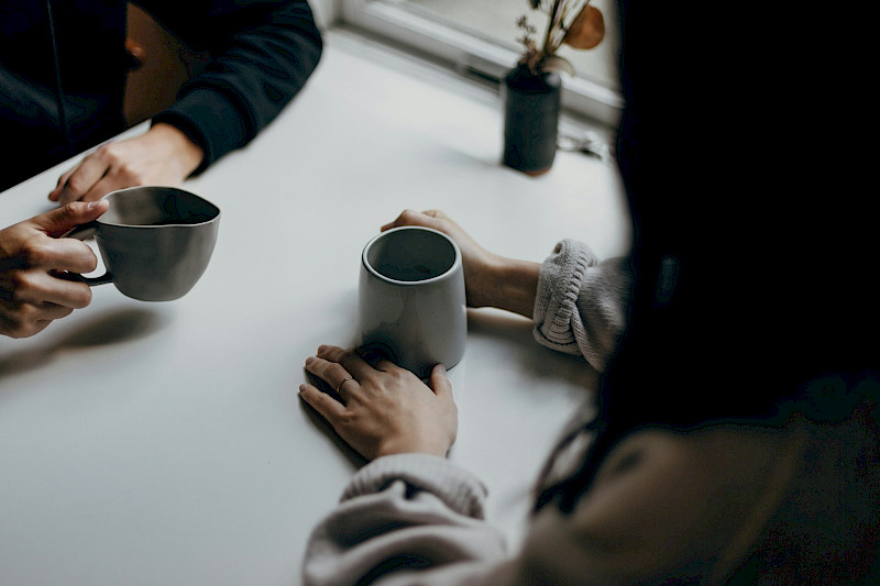Das Bild zeigt 4 Hände. 2 Personen sitzen sich gegenüber und haben jeweils graue Kaffeetassen mit Kaffee in der Hand. Neben den Personen ist ein Ausschnitt eines Fensters und eine Blumenvase mit Blumen zu sehen.