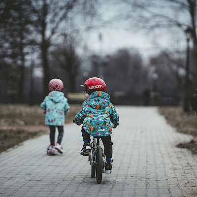 Das Bild zeigt zwei Kleinkinder, die auf einem Roller sowie auf einem Kinderfahrrad auf einem Weg unterwegs sind. Der Weg ist grau gepflastert. Neben dem Weg befinden sich Bäume. der Hintergrund zeigt graues, winterliches Wetter.