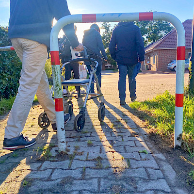 Das Bild zeigt mehrere Personen, die zu Fuß unterwegs sind. Die hintere Person ist auf einen Rollator angewiesen.  Der Hintergrund des Bildes erscheint sonnig, außerdem ist dort ein Haus in einer Wohnsiedlung zu sehen.