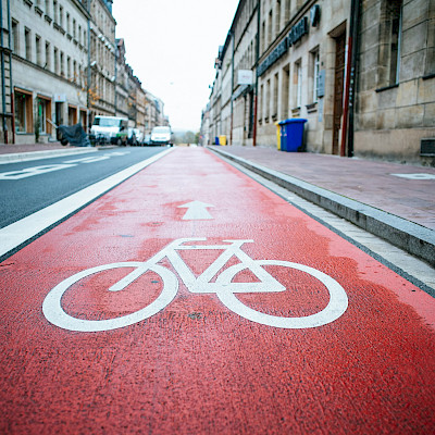 Das Bild zeigt ein Piktogramm, das eine Fahrradstraße markieren soll. Im Hintergrund sind parkende Autos und Häuser zu sehen.
