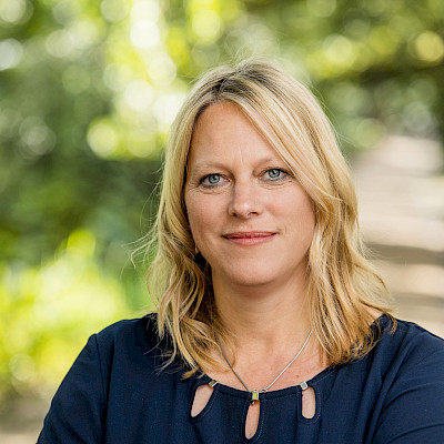 Das Bild zeigt  ein Portraitfoto von Dr. Maike Schaefer, der Senatorin a.D. für Klimaschutz, Umwelt, Mobilität, Stadtentwicklung und Wohnungsbau und Bürgermeisterin der Freien Hansestadt Bremen. Dr. Maike Schaefer trägt ein dunkelblaues Oberteil, ihre Haare sind hellblond. der Hintergrund des Portraitfotos erscheint sommerlich, es sind Umrisse von Bäu,men und blühenden Pflanzen zu sehen.