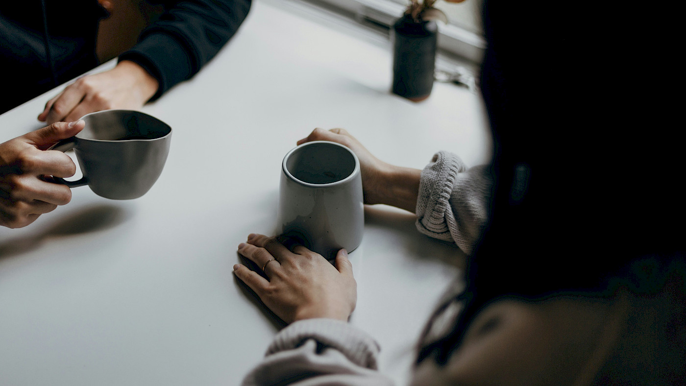 Das Bild zeigt 4 Hände. 2 Personen sitzen sich gegenüber und haben jeweils graue Kaffeetassen mit Kaffee in der Hand. Neben den Personen ist ein Ausschnitt eines Fensters und eine Blumenvase mit Blumen zu sehen.