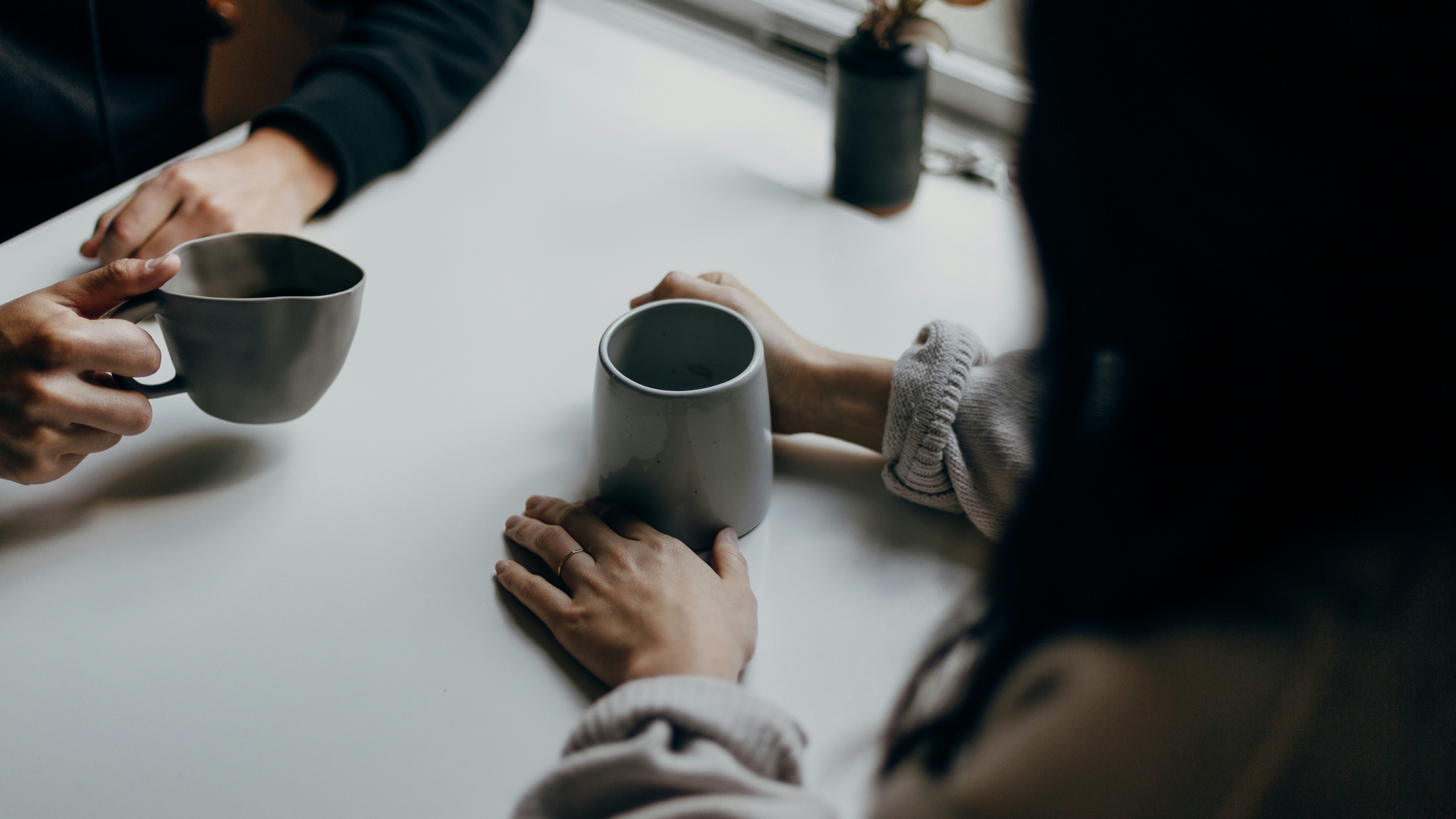 Das Bild zeigt 4 Hände. 2 Personen sitzen sich gegenüber und haben jeweils graue Kaffeetassen mit Kaffee in der Hand. Neben den Personen ist ein Ausschnitt eines Fensters und eine Blumenvase mit Blumen zu sehen.