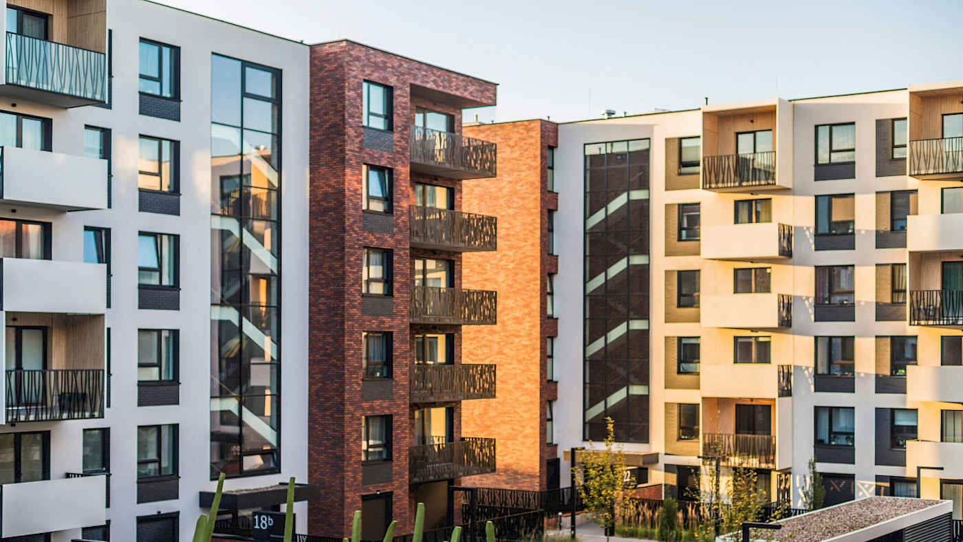 Das Bild zeigt ein Wohnquartier mit mehreren Mehrfamilienhäusern, die Balkone haben. Die Häuser sind weiß und rötlich. Im Vordergrund sind Grünflächen zu sehnen.