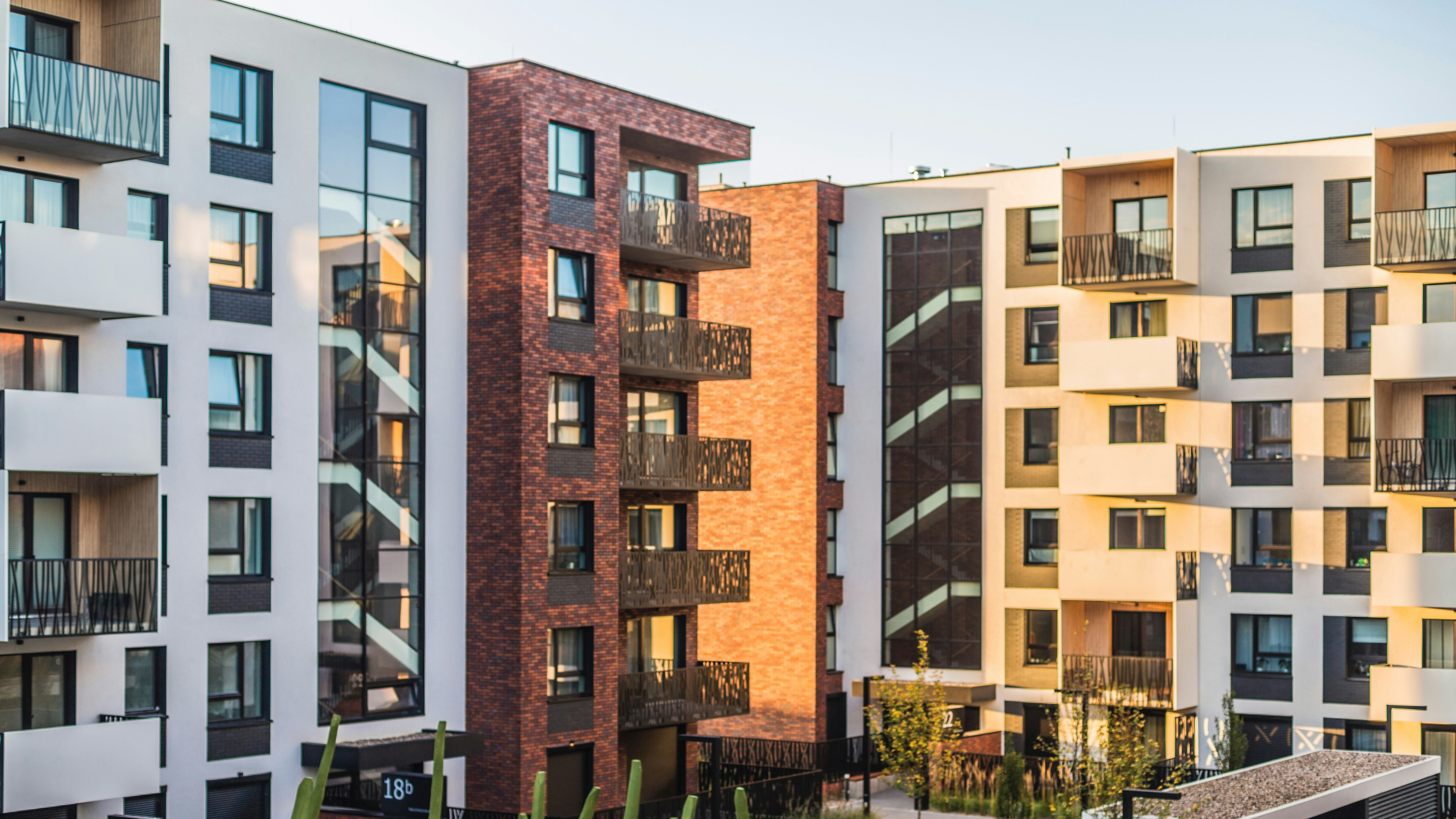 Das Bild zeigt ein Wohnquartier mit mehreren Mehrfamilienhäusern, die Balkone haben. Die Häuser sind weiß und rötlich. Im Vordergrund sind Grünflächen zu sehnen.