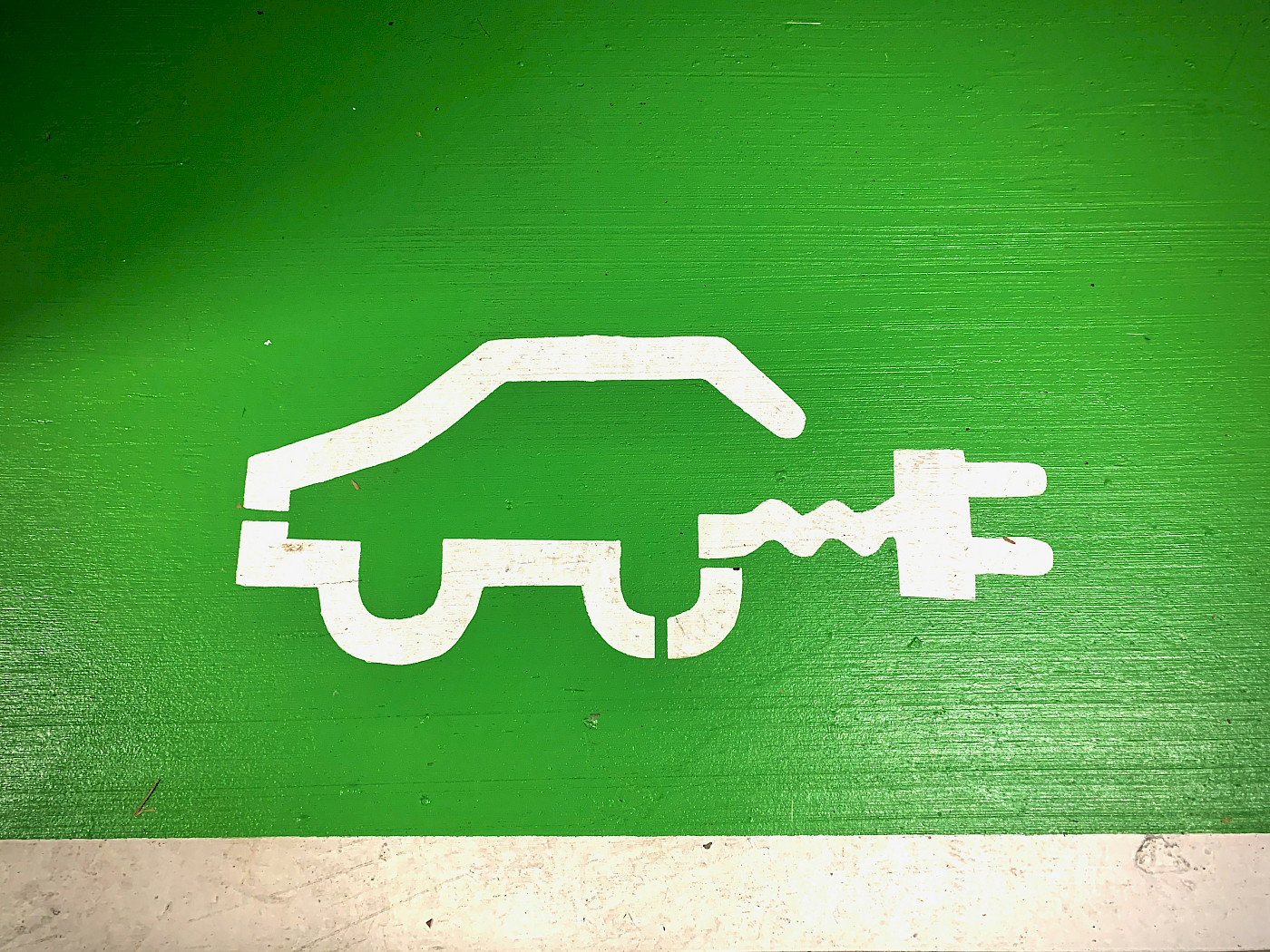 Es ist eine Ladestation für Elektroautos und der entsprechende Parkplatz dazu zu sehen. der Parkplatz ist durch ein großflächiges grünes Piktogramm mit weißem Auto gekennzeichnet. der Hintergrund des Fotos ist herbstlich, man sieht einen Baum und eine Hecke mit buntgefärbten Blättern.