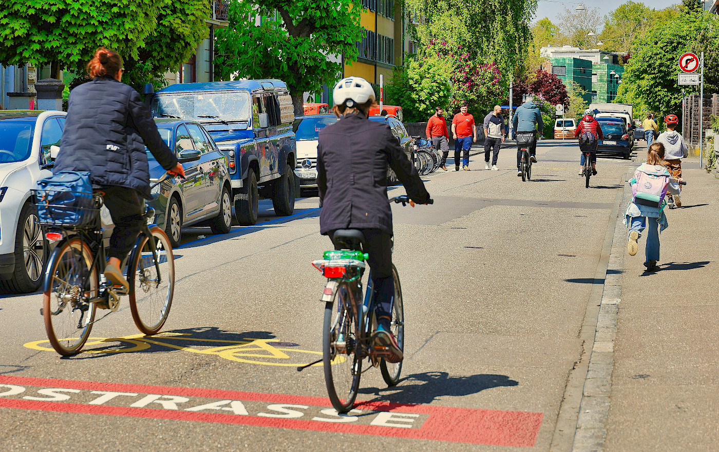 Auf dem Bild siind zwei Radfahrende von hinten zu erkennen, die sich auf einer Fahrradstraße befinden. Vor hinen sind weitere Radfahrende zu sehen und auch Zufußgehende.