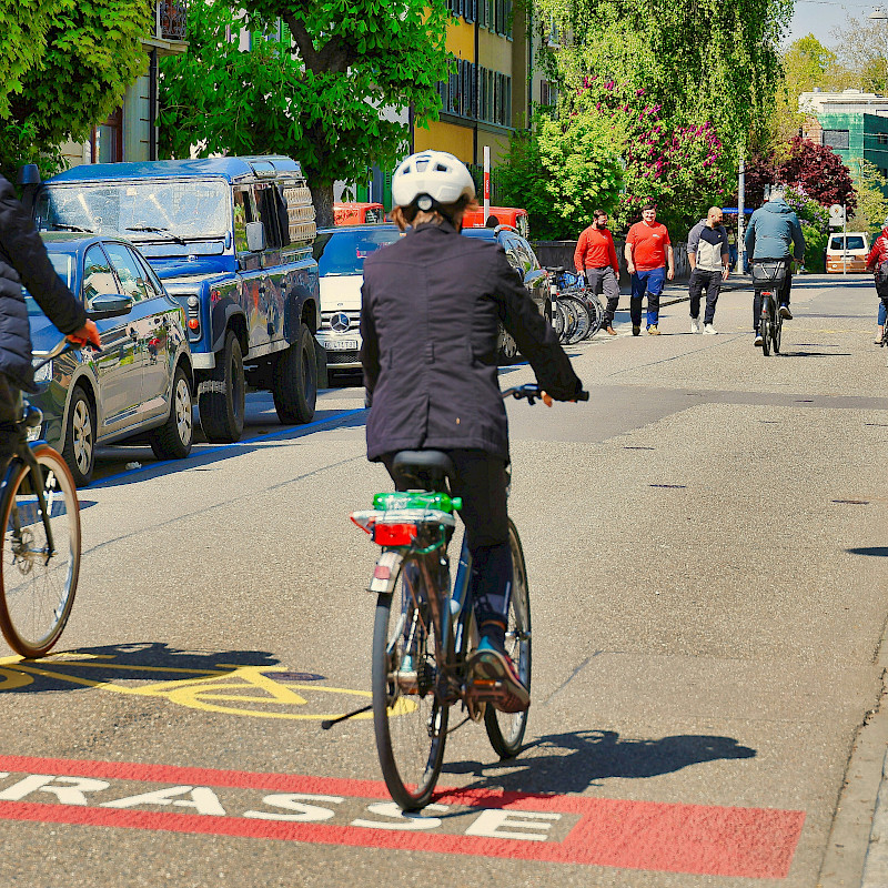 Auf dem Bild siind zwei Radfahrende von hinten zu erkennen, die sich auf einer Fahrradstraße befinden. Vor hinen sind weitere Radfahrende zu sehen und auch Zufußgehende.