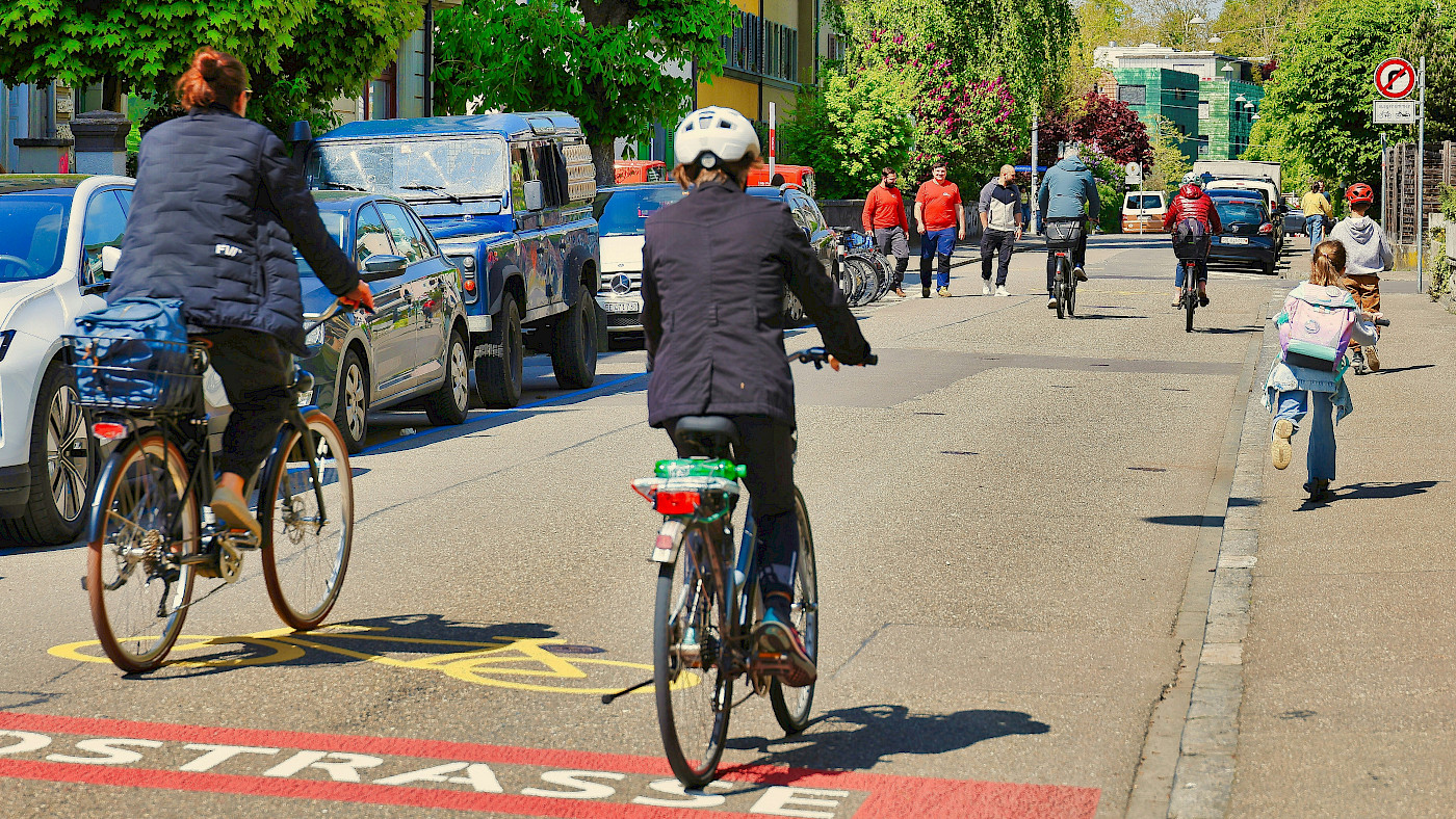 Auf dem Bild sind zwei Radfahrende von hinten zu erkennen, die sich auf einer Fahrradstraße befinden. Es sind weitere Radfahrende und auch Zufußgehend am Ende der Straße von hinten zusehene.