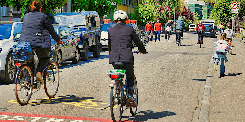 Auf dem Bild sind zwei Radfahrende von hinten zu erkennen, die sich auf einer Fahrradstraße befinden. Es sind weitere Radfahrende und auch Zufußgehend am Ende der Straße von hinten zusehene.