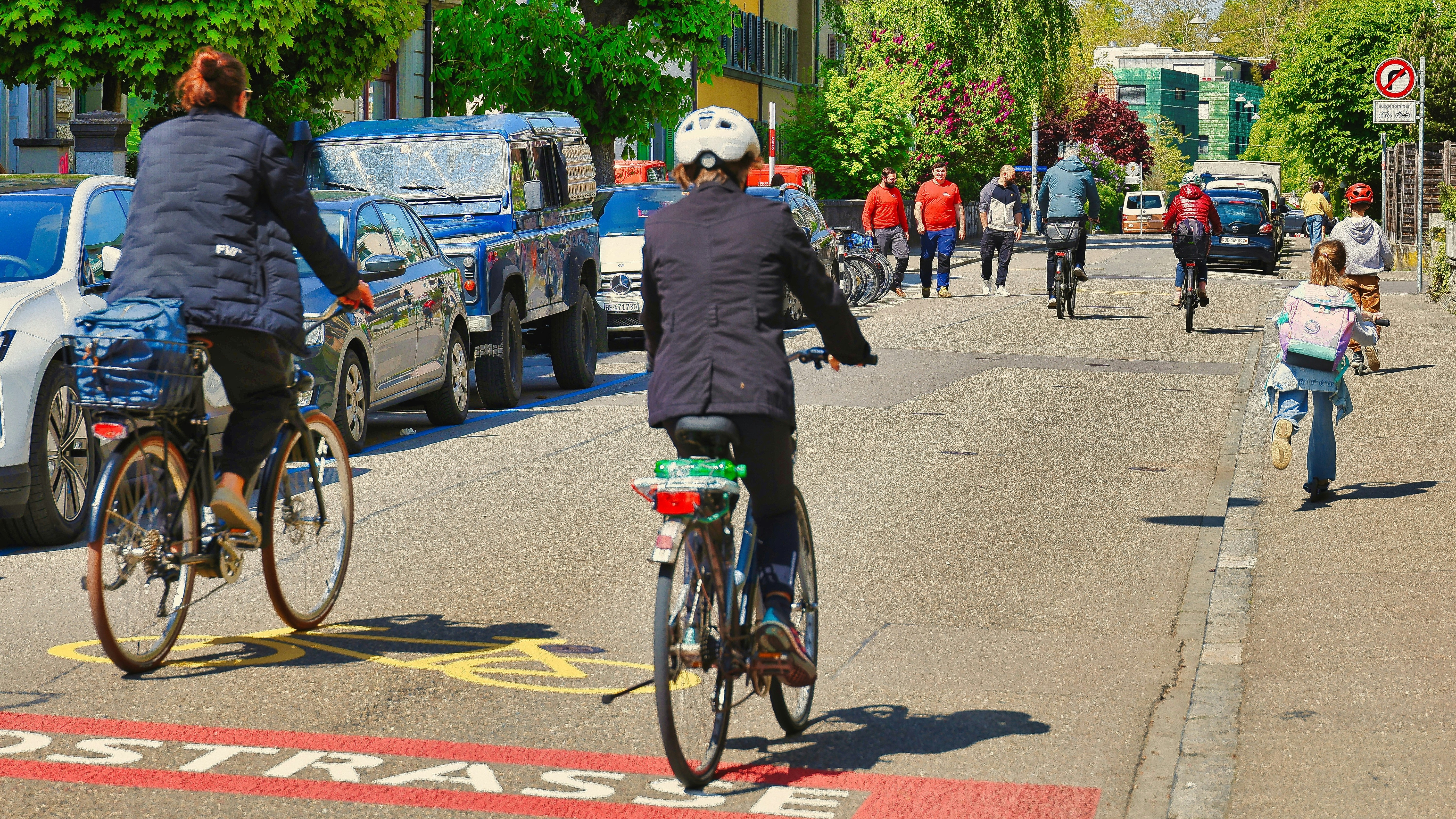 Auf dem Bild sind zwei Radfahrende von hinten zu erkennen, die sich auf einer Fahrradstraße befinden. Es sind weitere Radfahrende und auch Zufußgehend am Ende der Straße von hinten zusehene.