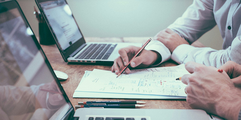 Auf dem Bild sind vier Hände von 2 Personen zu sehen. Die Personen haben Stift und Zettel vor sich liegen und sitzen an 2 Laptops, um dort zu arbeiten. Das Bild zeigt eine