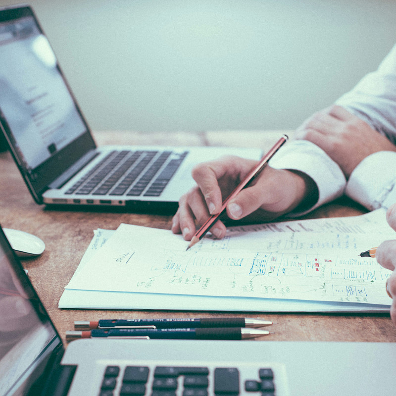 Auf dem Bild sind vier Hände von 2 Personen zu sehen. Die Personen haben Stift und Zettel vor sich liegen und sitzen an 2 Laptops, um dort zu arbeiten. Das Bild zeigt eine