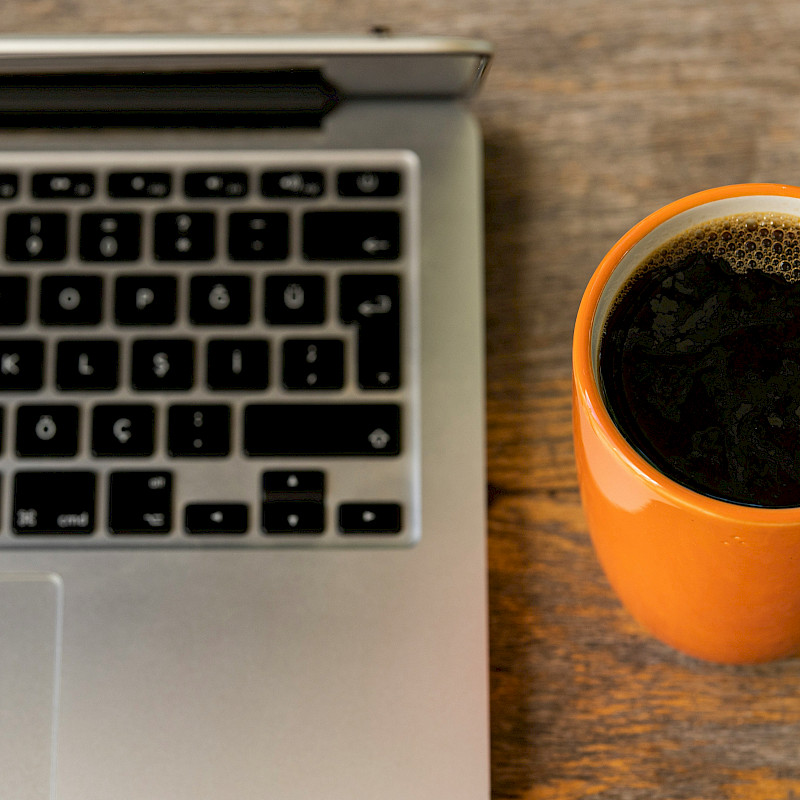 Auf dem Bild ist der AUsschnitt eines silbernen Laptops und dessen Tatstatur zu erkennen. Daneben sreht eine orangefarbige Tasse, die mit schwarzem Kaffee gefüllt ist. Der Laptop und die Tasse stehen auf einem massiven Holztisch. Der Untergrund des Tisches ist zu erkennen.