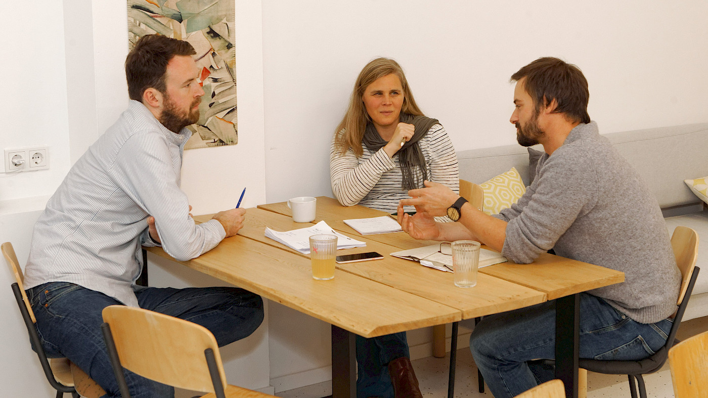 Das Bild zeigt drei Personen (zwei Männer mit braunen haaren und Bart sowie eine Frau mit langen Haaren), die sich an einem Tisch aus Holz gegenüber sitzen. Sie disktuieren gemeinam. Auf dem Tisch befinden sich Unterlagen, Schreibutensilien und Getränke.