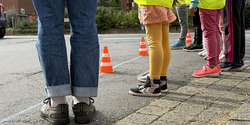 Das Bild zeigt die Füße von mindestens 5 Personen. Unter den Personen befinden sich auch Kinder, die bunte Kleidung tragen. Die Personen stehen auf einer Straße, vor ihnen befinden sich Verkehrspilonen, die etwas markieren. Der Ausschnitt des Bildes fokussiert die Füße der Personen.