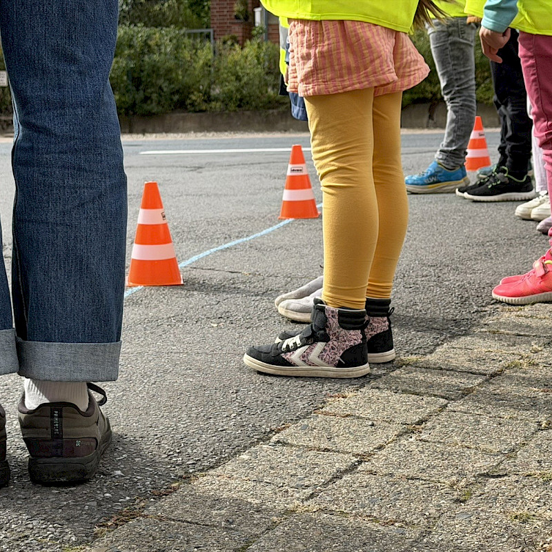 Das Bild zeigt die Füße von mindestens 5 Personen. Unter den Personen befinden sich auch Kinder, die bunte Kleidung tragen. Die Personen stehen auf einer Straße, vor ihnen befinden sich Verkehrspilonen, die etwas markieren. Der Ausschnitt des Bildes fokussiert die Füße der Personen.