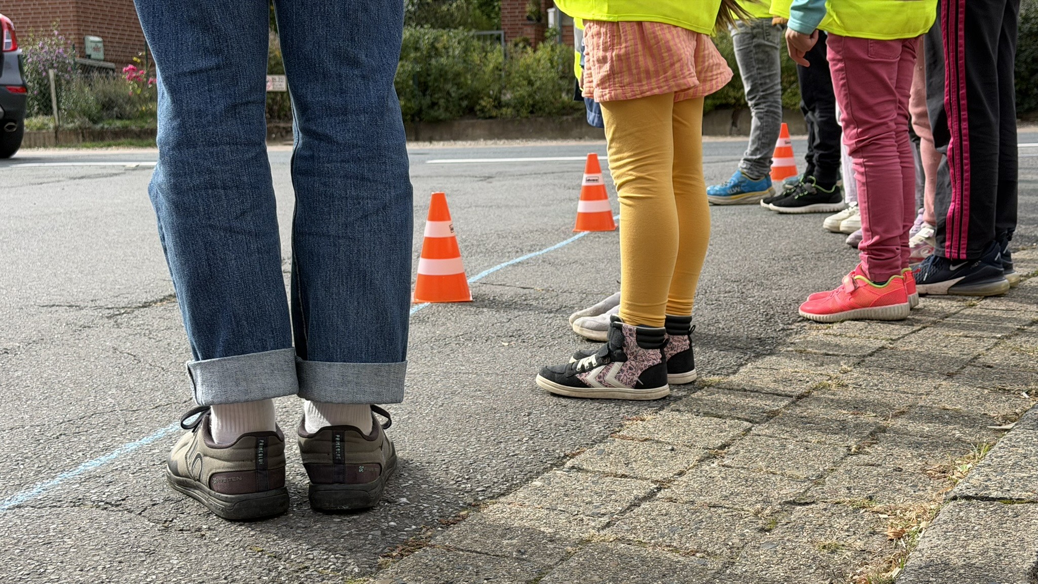 Das Bild zeigt die Füße von mindestens 5 Personen. Unter den Personen befinden sich auch Kinder, die bunte Kleidung tragen. Die Personen stehen auf einer Straße, vor ihnen befinden sich Verkehrspilonen, die etwas markieren. Der Ausschnitt des Bildes fokussiert die Füße der Personen.