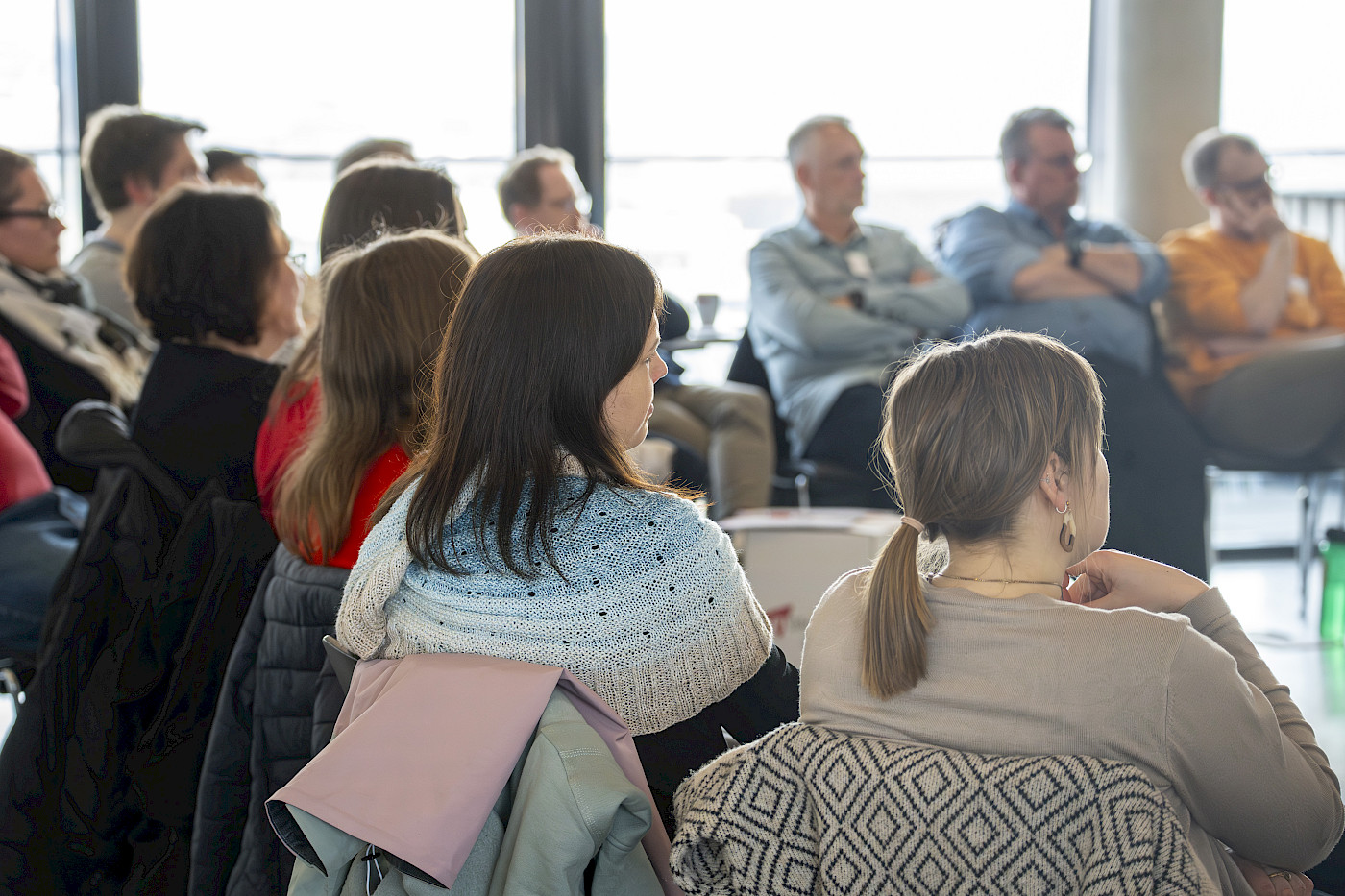 Das Bild zeigt mehrere Personen, die in einem Stuhlkreis zueinander sitzen und die einem Vortrag zuhören. die Personen sind lediglich von hinten zu erkennen.