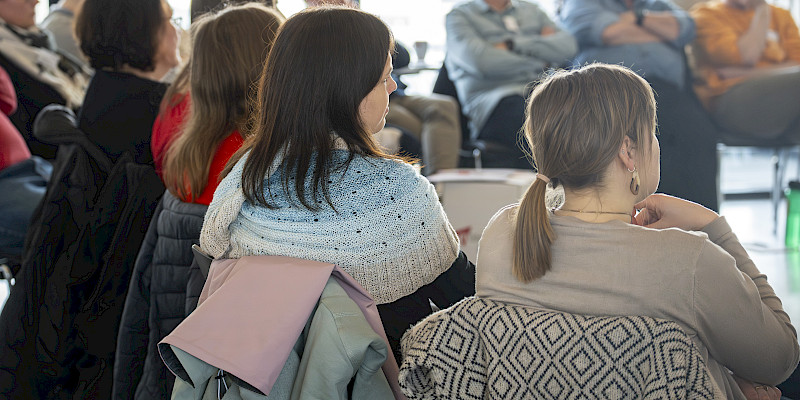 Das Bild zeigt mehrere Personen, die in einem Stuhlkreis zueinander sitzen und die einem Vortrag zuhören. die Personen sind lediglich von hinten zu erkennen.