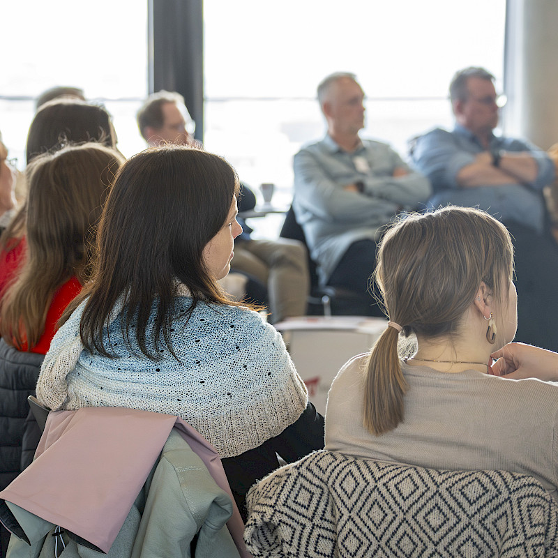 Das Bild zeigt mehrere Personen, die in einem Stuhlkreis zueinander sitzen und die einem Vortrag zuhören. die Personen sind lediglich von hinten zu erkennen.
