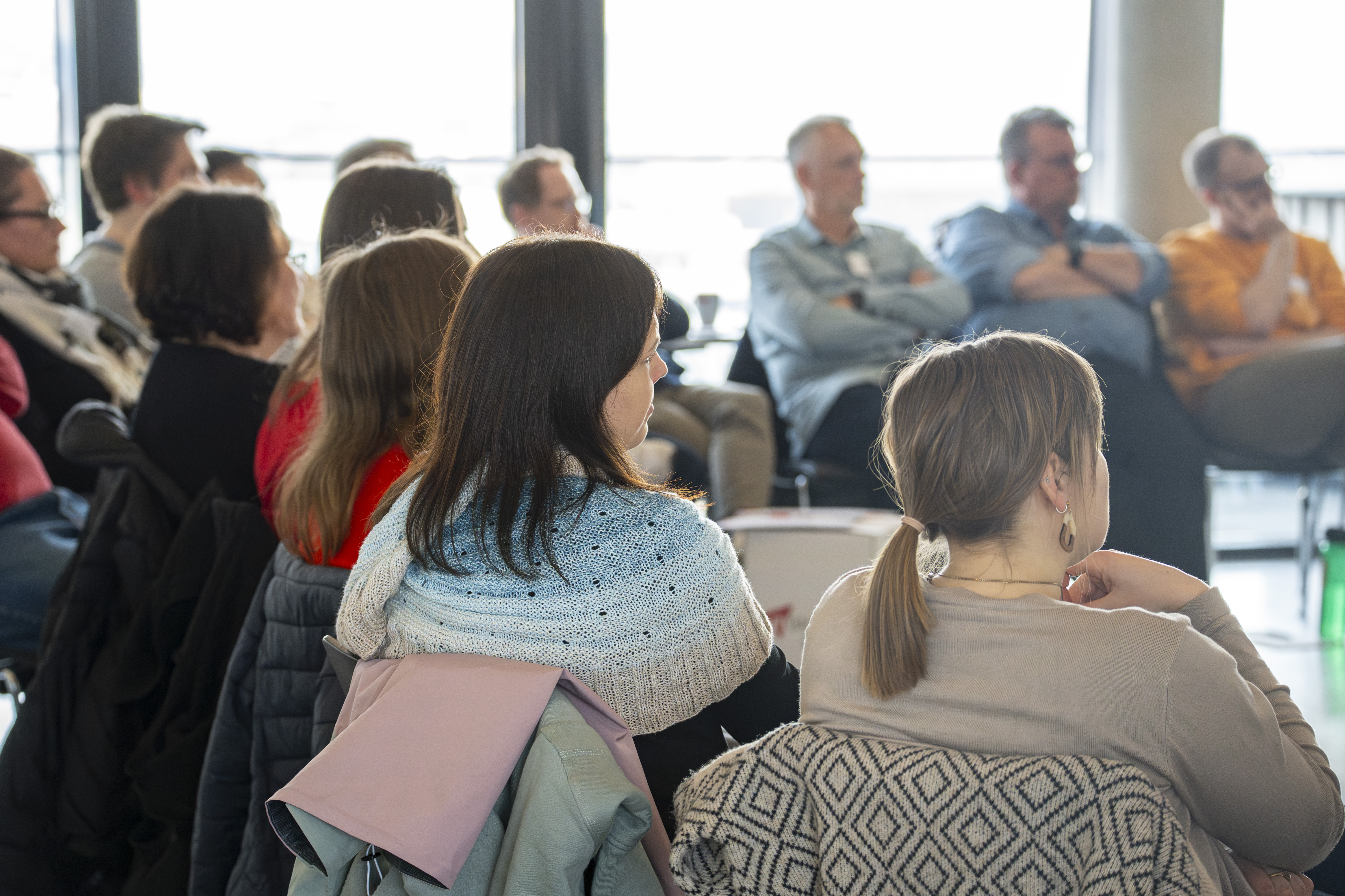 Das Bild zeigt mehrere Personen, die in einem Stuhlkreis zueinander sitzen und die einem Vortrag zuhören. die Personen sind lediglich von hinten zu erkennen.
