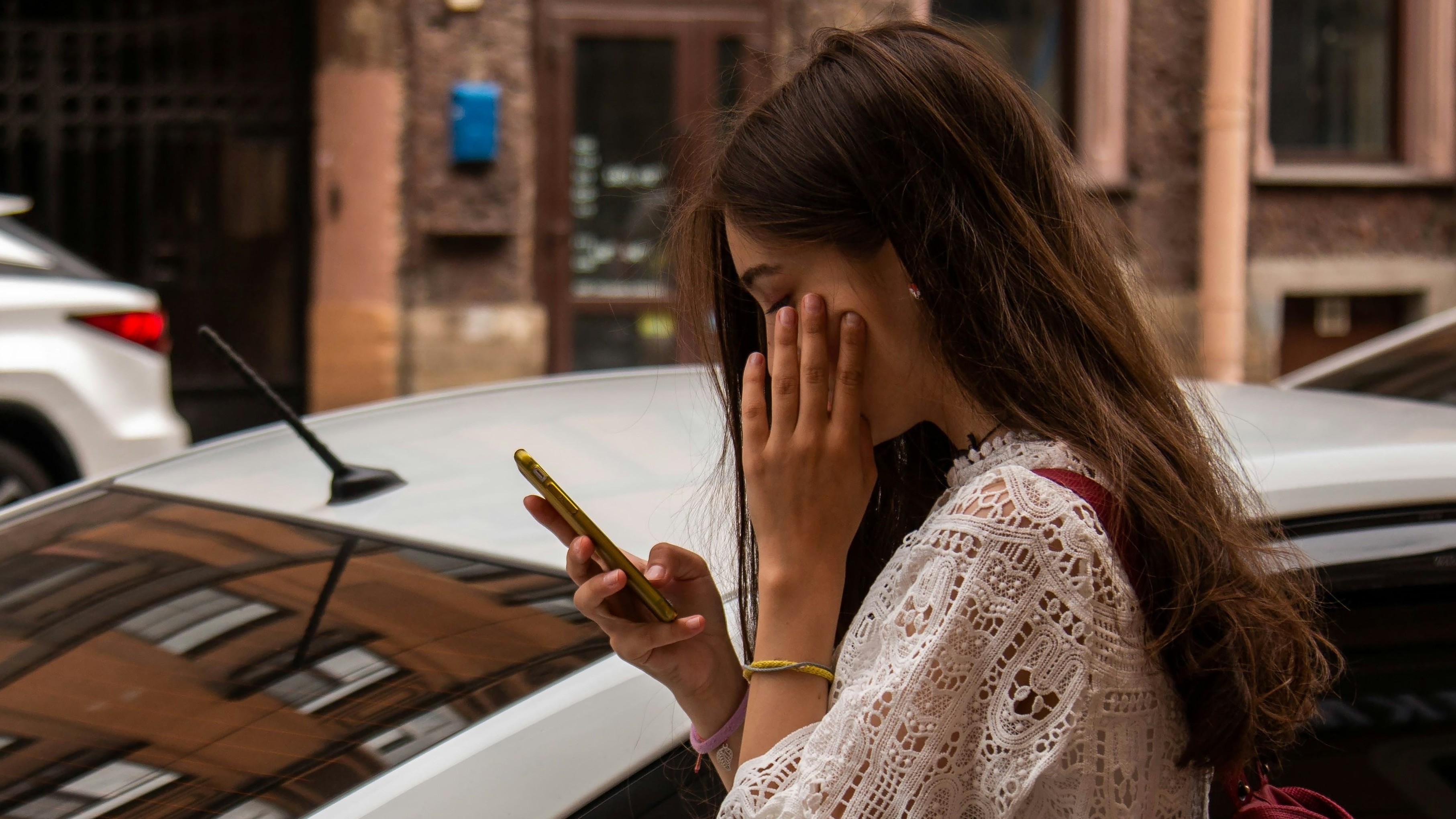 Das Bild zeigt eine junge Frau, die am Handy ist und die sich hinter einem weißen Auto befindet. Sie scheint zu warten. Gegenüber ist ein weiteres farbiges Auto zu sehen.