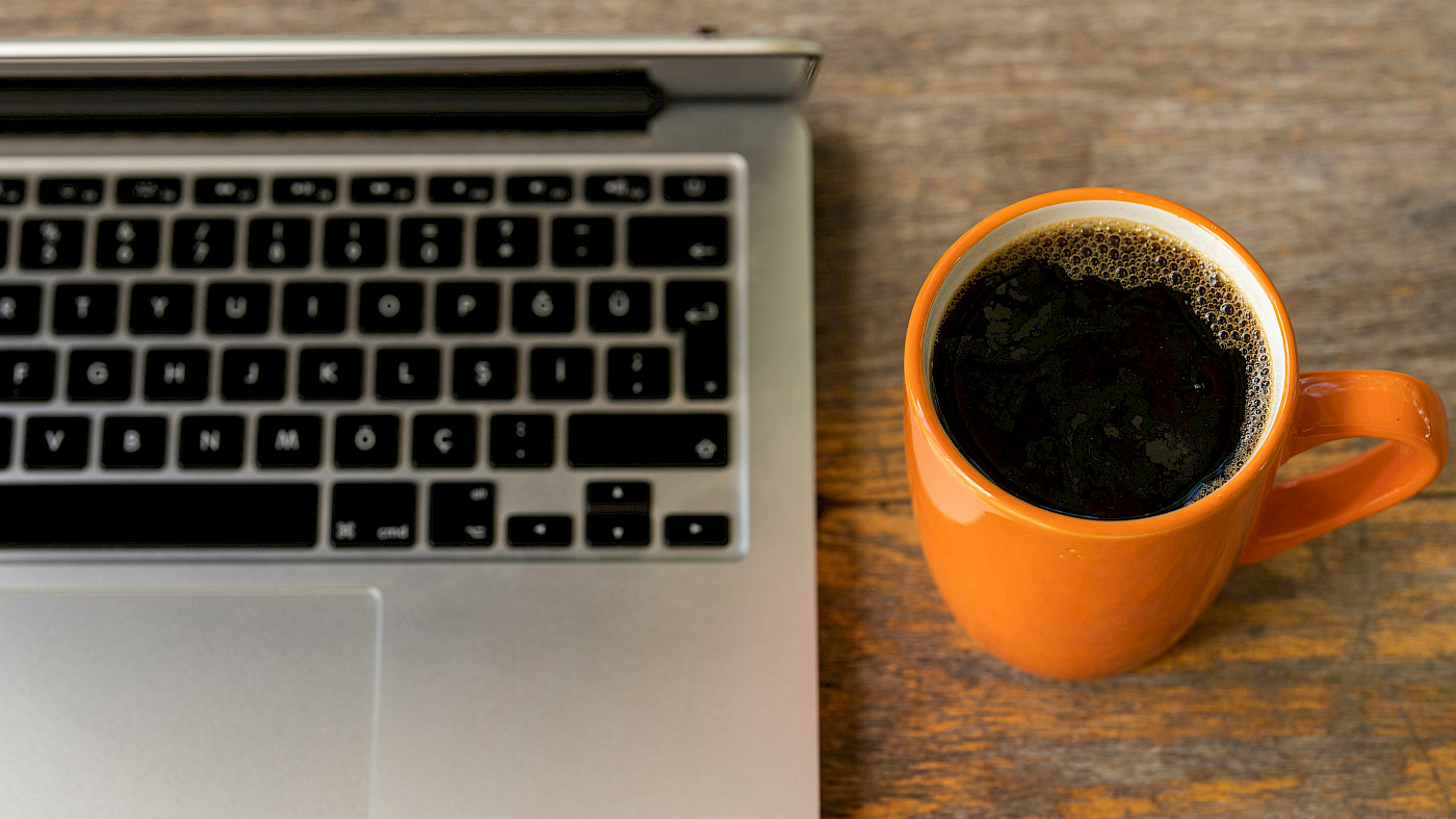 Auf dem Bild ist der AUsschnitt eines silbernen Laptops und dessen Tatstatur zu erkennen. Daneben sreht eine orangefarbige Tasse, die mit schwarzem Kaffee gefüllt ist. Der Laptop und die Tasse stehen auf einem massiven Holztisch. Der Untergrund des Tisches ist zu erkennen.