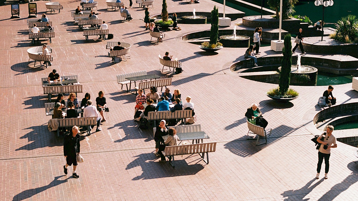 Auf dem Bild ist ein Shared Space zu sehen. Viele Menschen sitzen auf einem Platz auf Bänken und Sitzmöbeln, daneben laufen Menschen über den Platz. Es ist viel Grün und Beschattung um diesen Platz.