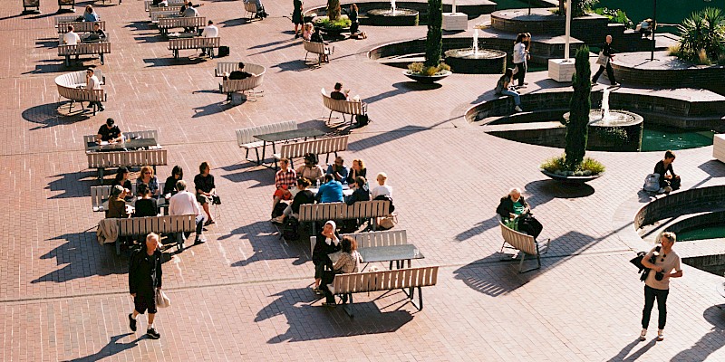 Auf dem Bild ist ein Shared Space zu sehen. Viele Menschen sitzen auf einem Platz auf Bänken und Sitzmöbeln, daneben laufen Menschen über den Platz. Es ist viel Grün und Beschattung um diesen Platz.