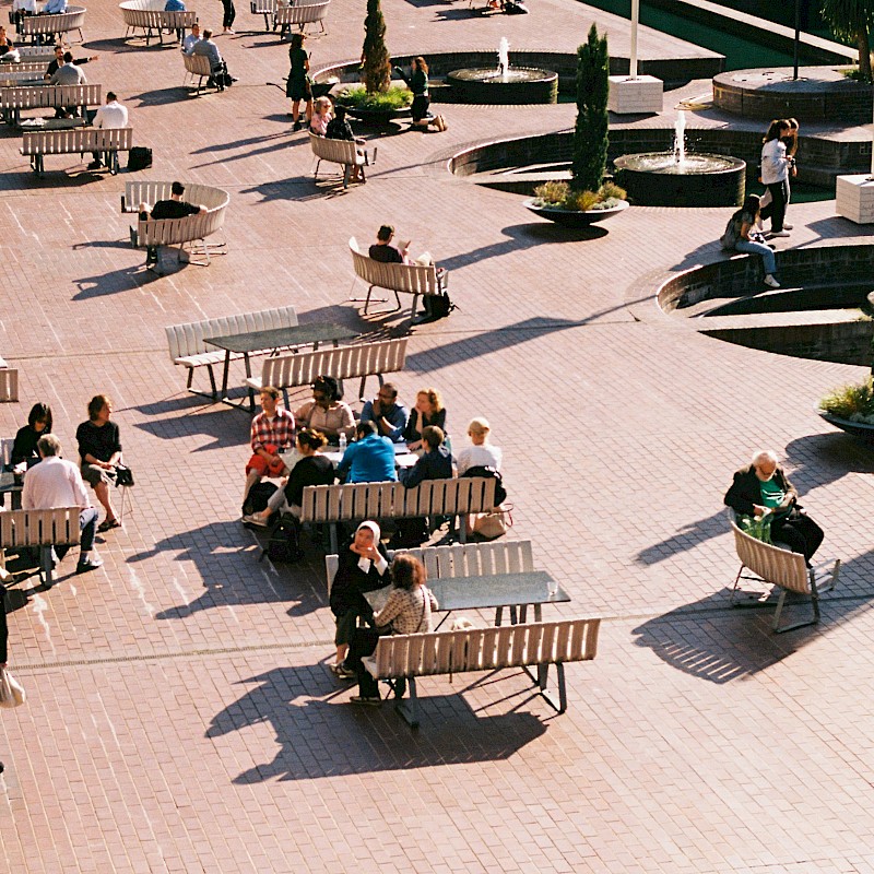Auf dem Bild ist ein Shared Space zu sehen. Viele Menschen sitzen auf einem Platz auf Bänken und Sitzmöbeln, daneben laufen Menschen über den Platz. Es ist viel Grün und Beschattung um diesen Platz.