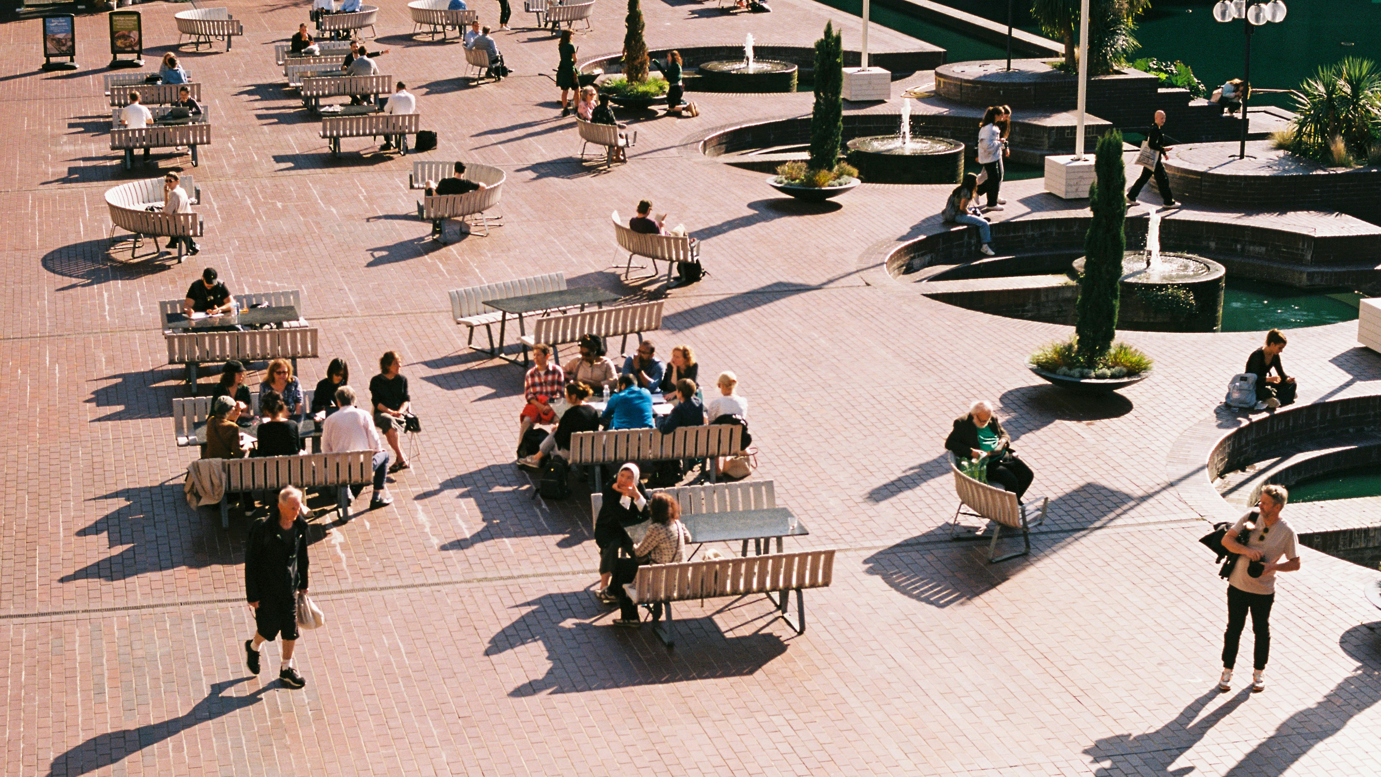 Auf dem Bild ist ein Shared Space zu sehen. Viele Menschen sitzen auf einem Platz auf Bänken und Sitzmöbeln, daneben laufen Menschen über den Platz. Es ist viel Grün und Beschattung um diesen Platz.