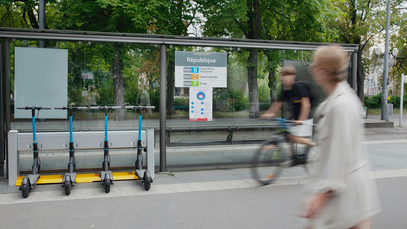 Auf dem Bild ist eine Bushaltestelle zu sehen. An dieser Bushatestelle sind auch E-Roller abgestellt. Außerdem ist ein Fahrradfahrer zu sehen. Im Vordergrund ist eine Fußgängerin zu sehen.