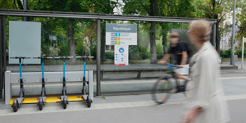 Auf dem Bild ist eine Bushaltestelle zu sehen. An dieser Bushatestelle sind auch E-Roller abgestellt. Außerdem ist ein Fahrradfahrer zu sehen. Im Vordergrund ist eine Fußgängerin zu sehen.