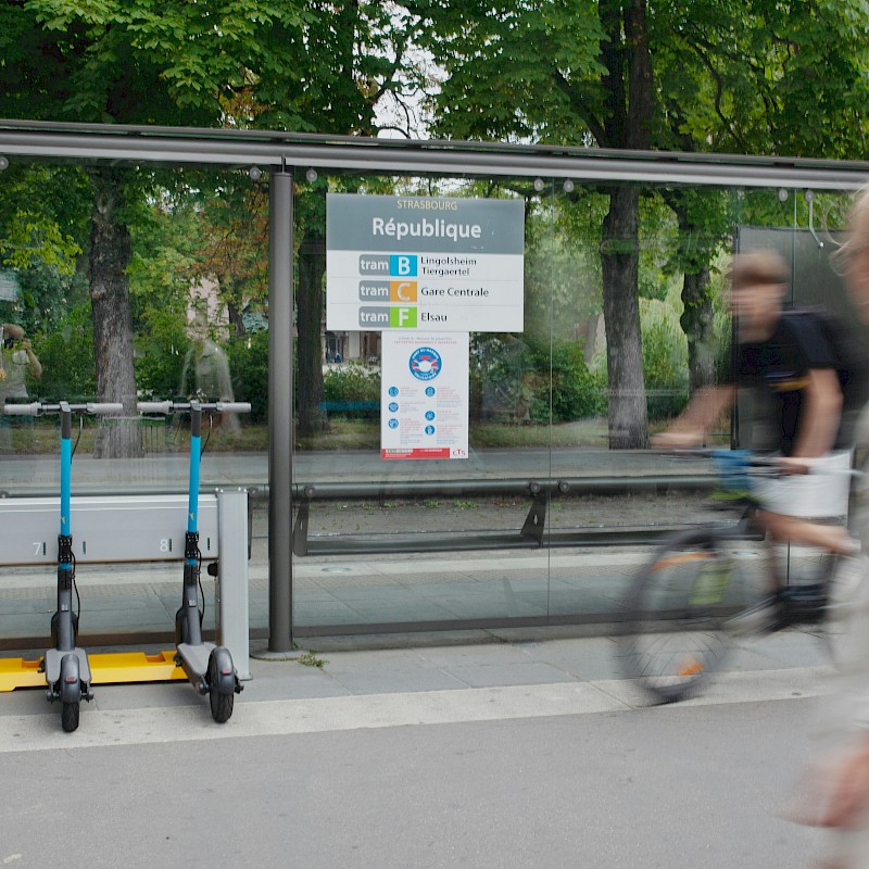 Auf dem Bild ist eine Bushaltestelle zu sehen. An dieser Bushatestelle sind auch E-Roller abgestellt. Außerdem ist ein Fahrradfahrer zu sehen. Im Vordergrund ist eine Fußgängerin zu sehen.