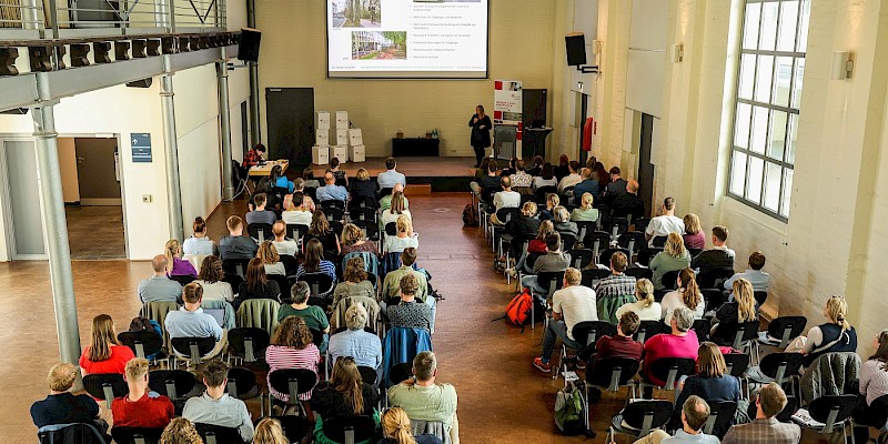 Das Bild zeigt eine Veranstaltung von oben. Es sind Stuhlreihen zu sehen, auf den Stühlen sitzen Menschen. Zwischen den beiden Stuhlreihen ist ein Gang. Das Publikum schaut nach vorne, vorne auf der Bühne ist eine Referentin zu sehen. vor ihr ist eine Leinwand abgebildet, die die Präsentation der Referentin zeigt.