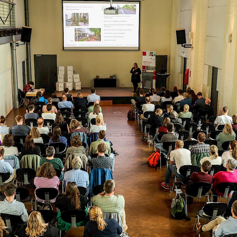 Das Bild zeigt eine Veranstaltung von oben. Es sind Stuhlreihen zu sehen, auf den Stühlen sitzen Menschen. Zwischen den beiden Stuhlreihen ist ein Gang. Das Publikum schaut nach vorne, vorne auf der Bühne ist eine Referentin zu sehen. vor ihr ist eine Leinwand abgebildet, die die Präsentation der Referentin zeigt.