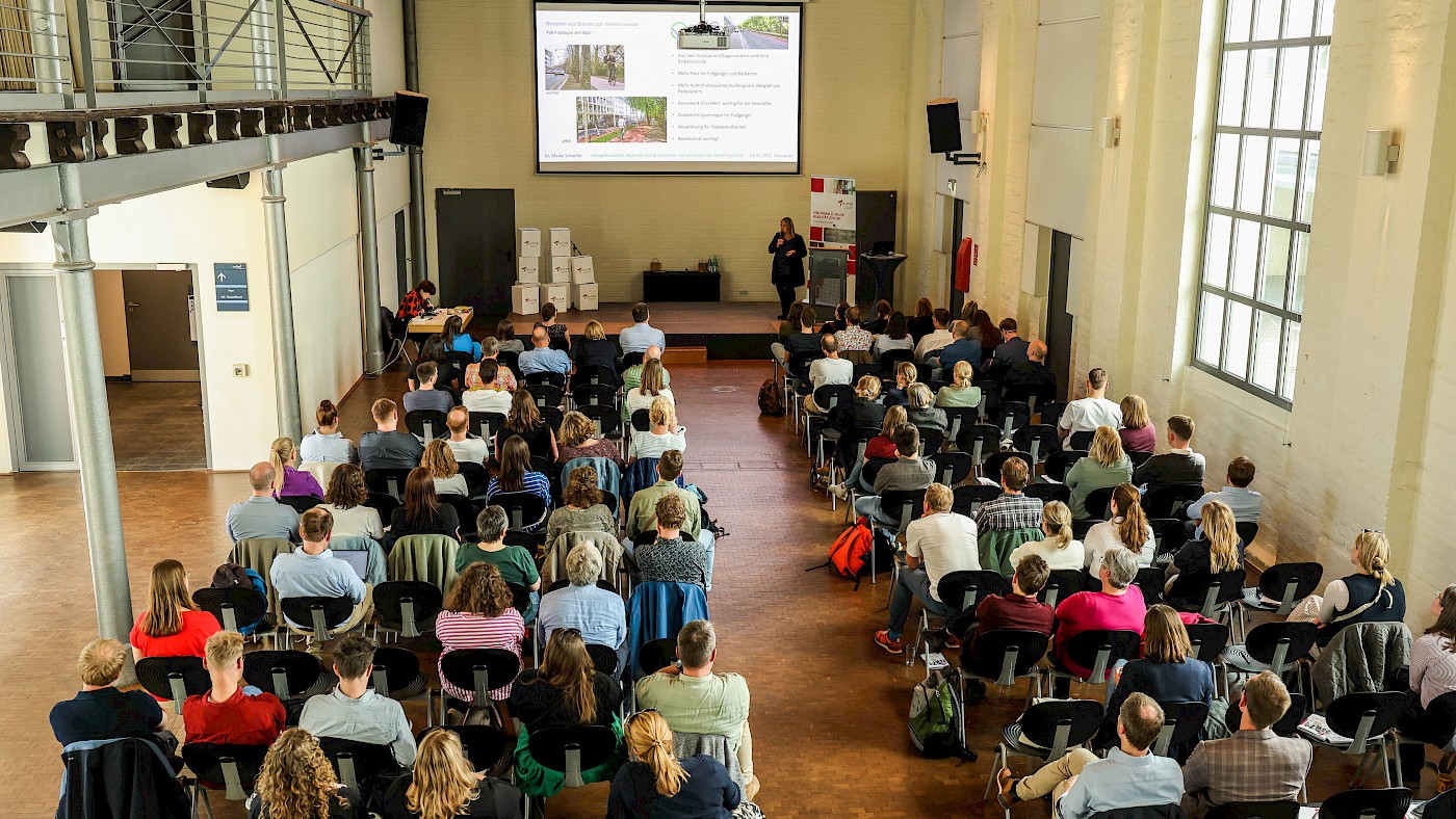 Das Bild zeigt eine Veranstaltung von oben. Es sind Stuhlreihen zu sehen, auf den Stühlen sitzen Menschen. Zwischen den beiden Stuhlreihen ist ein Gang. Das Publikum schaut nach vorne, vorne auf der Bühne ist eine Referentin zu sehen. vor ihr ist eine Leinwand abgebildet, die die Präsentation der Referentin zeigt.