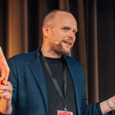 Das Bild zeigt ein Foto von Norbert Krause von krauses Projektdesign,der auf einer Bühne steht. In der rechten Hand hält er Moderationskarten.